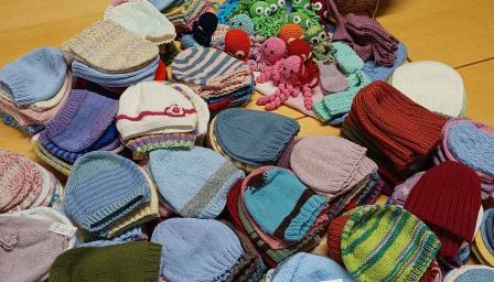 Pöydälle on aseteltu suuri määrä värikkäitä, käsin neulottuja vauvanmyssyjä siisteissä pinoissa. Yläosassa näkyy lisäksi virkattuja värikkäitä hahmoja, kuten mustekaloja ja eläimiä, sekä sukkia ja lapasia.