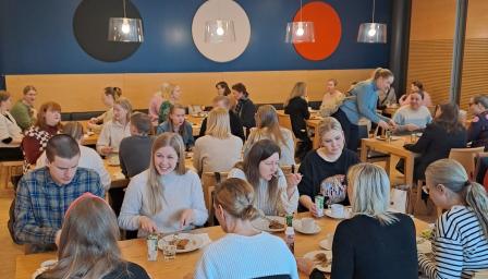 Ravintolasali täynnä ihmisiä syömässä ja seurustelemassa pitkien puupöytien ääressä. Taustalla näkyy sininen seinä, jossa on kolme suurta ympyrää väreissä musta, valkoinen ja punainen.