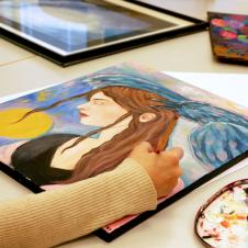 Maalaus, jossa on pitkähiuksinen nainen ja lintu. Käsi pitelee sivellintä maalauksen yllä.