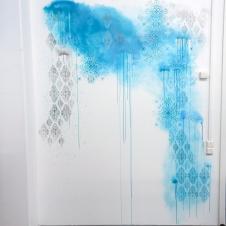 Seinällä näkyy abstrakti sinisävyinen maalaus, jossa yhdistyy sinistä spraymaalia ja koristeellisia sabluunakuvioita.