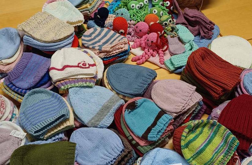Pöydälle on aseteltu suuri määrä värikkäitä, käsin neulottuja vauvanmyssyjä siisteissä pinoissa. Yläosassa näkyy lisäksi virkattuja värikkäitä hahmoja, kuten mustekaloja ja eläimiä, sekä sukkia ja lapasia.
