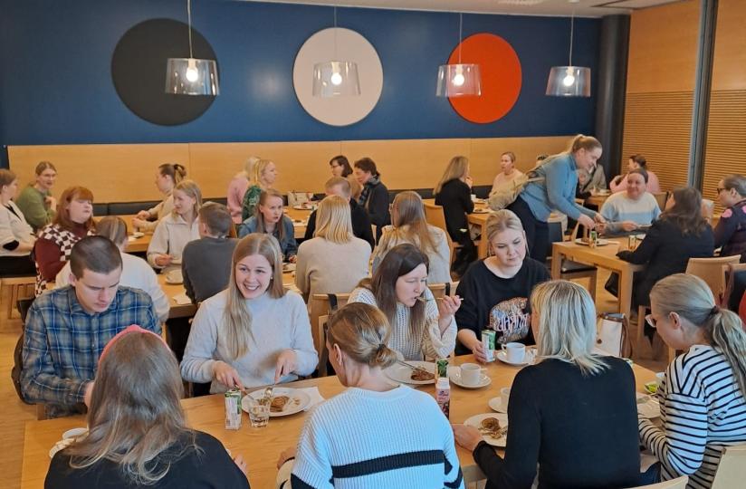 Ravintolasali täynnä ihmisiä syömässä ja seurustelemassa pitkien puupöytien ääressä. Taustalla näkyy sininen seinä, jossa on kolme suurta ympyrää väreissä musta, valkoinen ja punainen.