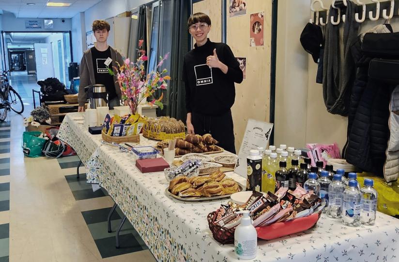Kahvilapöytä kuvattuna sivulta. Pöydän ääressä seisoo kaksi opiskelijaa, toinen antaa peukun merkiksi. Tarjoilut sisältävät pullia, suklaata ja juomia.