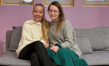 Maija ja Tytti Hultman istuvat harmaalla sohvalla vaaleanviolettia seinää vasten. Vasemmanpuoleinen nainen on Maija Hultman. Hän on pukeutunut valkoiseen neuleeseen ja mustiin housuihin, ja hänen pitkät vaaleat hiuksensa ovat toisella olkapäällä. Oikeanpuoleinen nainen on Tytti Hultman. Hän on pukeutunut harmaaseen neuleeseen ja vihreään hameeseen. Molemmat hymyilevät ja katsovat kameraan.