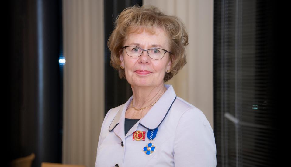 Sirkka Wiman katsoo kameraan. Hänellä on takin rinnuksessa kiinni punainen Kuntaliiton ansiomerkki, jossa on kultainen K-kirjain, sekä sininen AMKEn kultainen ansioristi.
