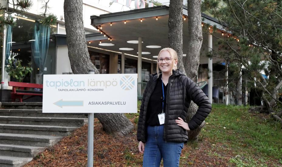 Emma Lahtinen seisoo rakennuksen edessä vieressään kyltti, jossa lukee Tapiolan Lämpö -konserni, asiakaspalvelu.