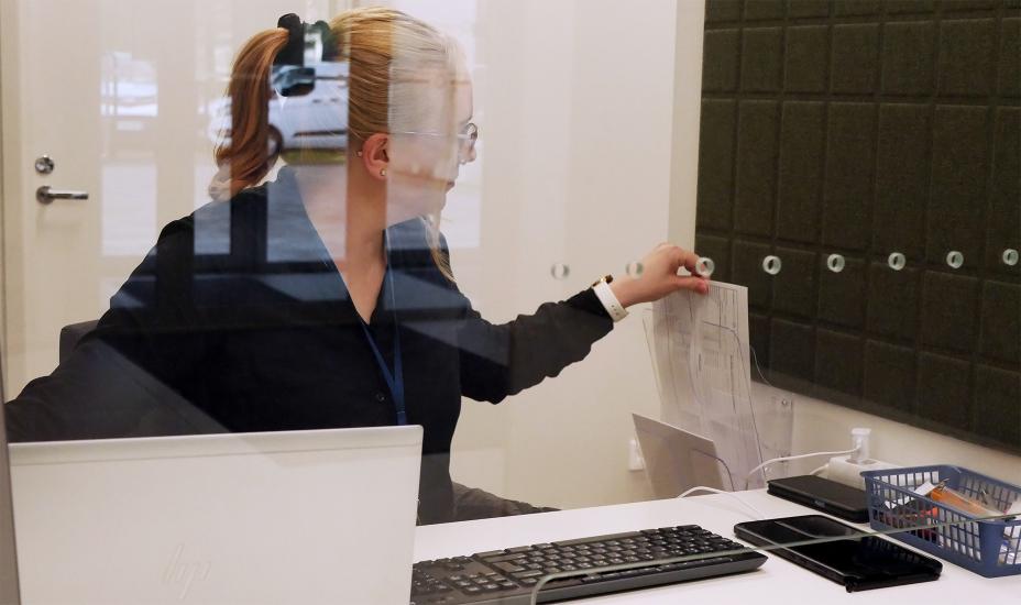 Emma Lahtinen istuu pöydän takana. Hänellä on edessään tietokone ja näppäimistö ja hän ottaa seinällä olevasta telineestä paperin, jossa lukee avaintenluovutus.