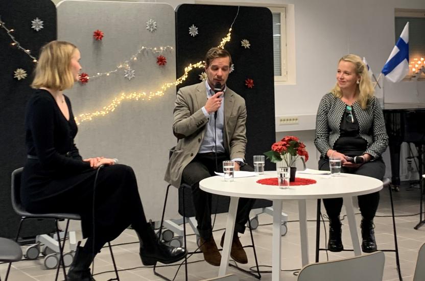 Kolme henkilöä istuu lavalla keskustelutilaisuudessa mikrofonien kanssa, taustalla harmaita ja mustia sermejä, joissa on valoja ja koristeita. Etualalla valkoinen pöytä, jossa joulutähti, laseja ja punainen liina; oikeassa reunassa näkyy Suomen lippu ja kynttilöitä ikkunalaudalla.