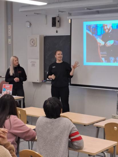 Kaksi henkilöä pitää esitystä luokassa valkokankaan edessä, ja taustalla näkyy kuva hymyilevästä baarityöntekijästä. Oppilaat seuraavat esitystä pulpettien äärestä keskittyneesti.