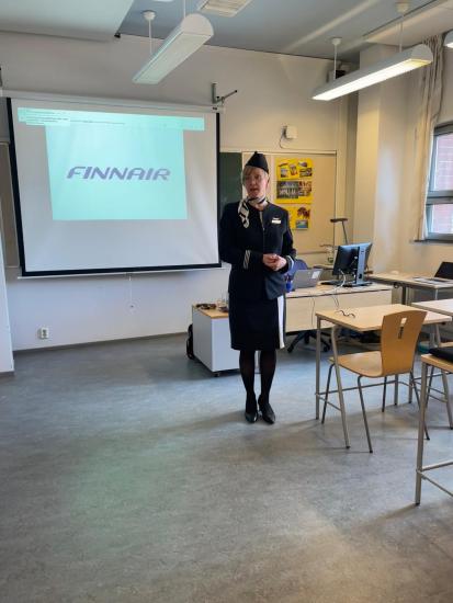 Finnairin lentoemännäksi pukeutunut henkilö seisoo luokan edessä esittelemässä, taustalla valkokankaalla näkyy Finnairin logo. Luokkatilassa on tietokone, tuoleja ja esitystarvikkeita.