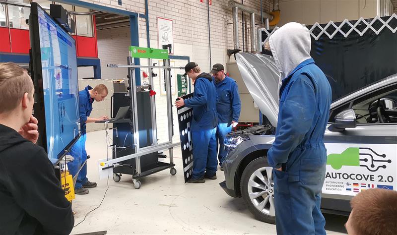 Autoalan opiskelijoita työhaalareissa tutkimassa auton teknisiä järjestelmiä korjaamoympäristössä. Etualalla näkyvässä autossa on "AUTOCOVE 2.0" -logo ja konepelti auki huoltoa varten.