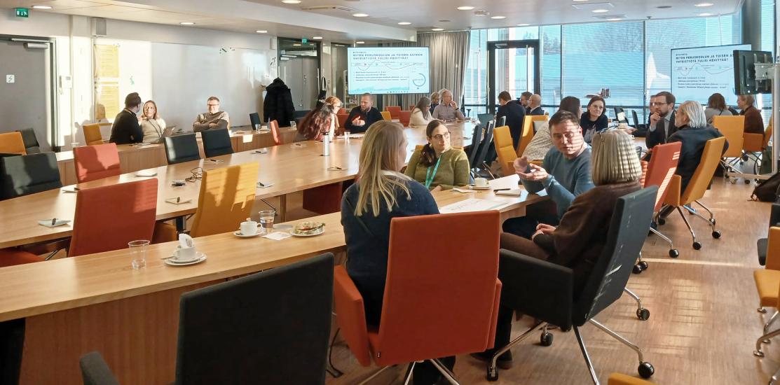 Kokoushuoneessa käynnissä työpajakeskustelu; osallistujat istuvat pöytien ääressä pienryhmissä ja taustalla näkyy esitysdia.