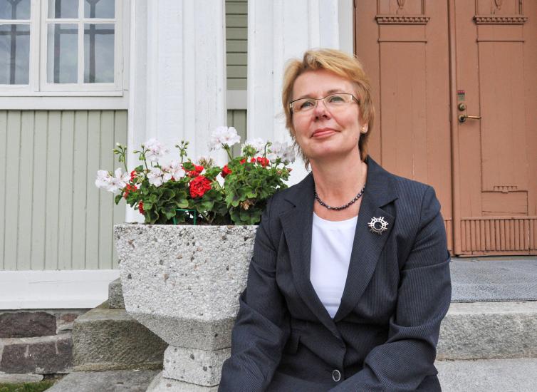 Sirkka Wiman istuu vanhan talon rappusilla. Vieressä on kesäkukkia ruukussa.
