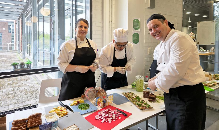 Kolme kokkiasuista henkilöä valmistaa tarjoiluleipiä pöydän ääressä. Etualalla näkyy retiisejä, yrttejä ja ruisleipää sekä leiville levitettävää vihreää tahnaa. Kaikki näyttävät keskittyneiltä ja hyväntuulisilta. Taustalla näkyy ikkunan kautta ulos sateiselle sisäpihalle.