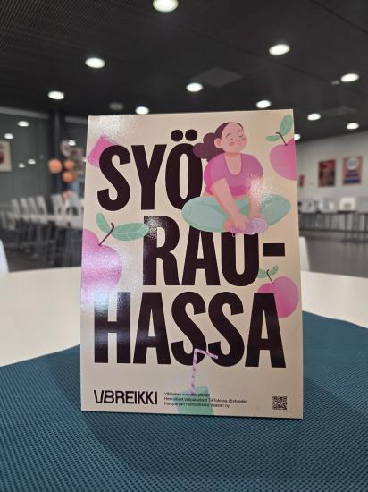 Pöydällä sinisen tabletin päällä on pystyssä oleva kampanjajuliste, jossa lukee isolla “Syö rauhassa”. Julisteessa on myös piirros meditoivasta hahmosta sekä vaaleanpunaisia hedelmäkuvituksia. Taustalla näkyy ruokasali, jossa on tyhjiä valkoisia tuoleja ja pöytiä.