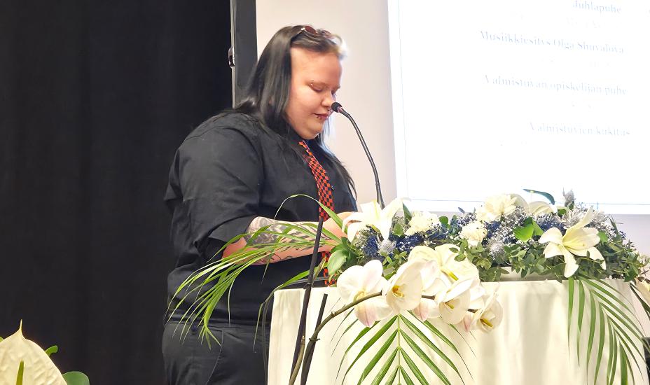 Kai Summala seisoo lavalla puhujankorokkeen takana ja katsoo paperiin. Edessä on kukkia.